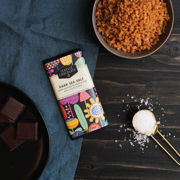 Seattle Chocolate Dark Sea Salt Dark Chocolate Truffle Bar with crunchy toffee next to chocolate, toffee, and salt on a table