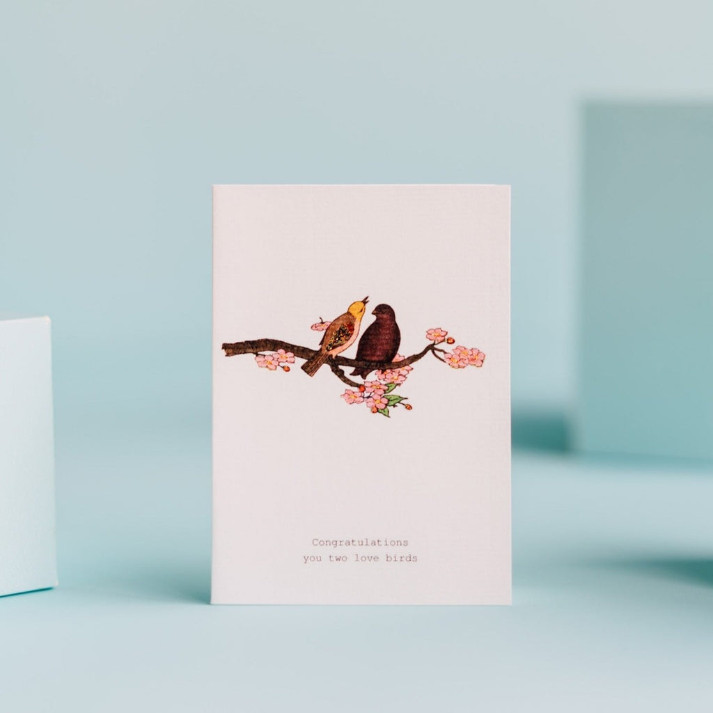 Greeting card reads: "Congratulations you two love birds" with a glittery illustration of two birds on a blossoming tree branch.
